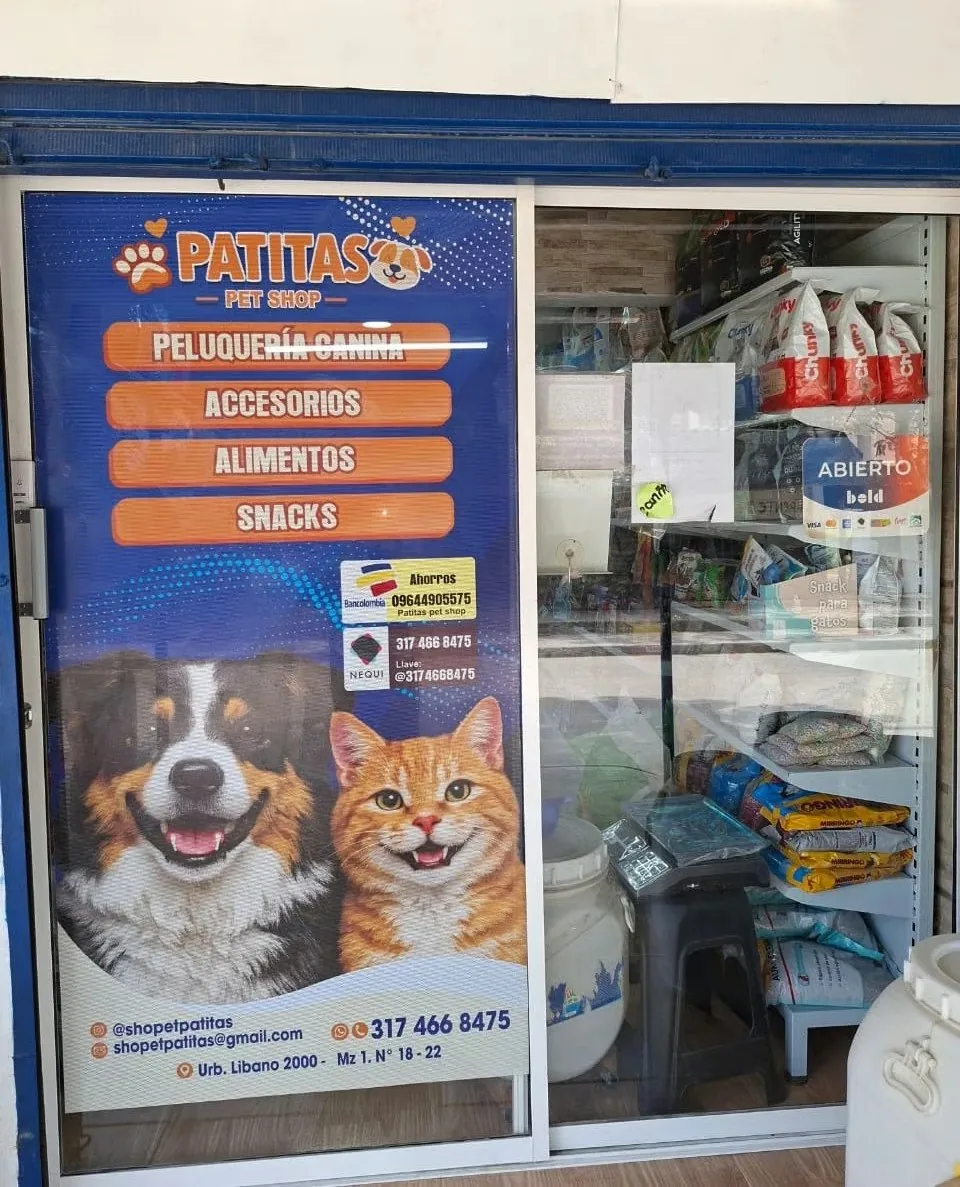 Fachada de Patitas Pet Shop con aviso de Peluquería Canina, Accesorios, Alimentos y Snacks, ilustraciones de un perro y un gato, y los estantes del local vistos por la puerta de vidrio